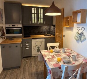 A compact kitchen with dark cabinets, a stone backsplash, a wooden countertop, and a dining area with a table set for two.
