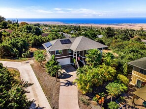 A single-story house with a garage, surrounded by lush greenery and a view of the ocean. - HALE LIO KAI: GORGEOUS HOME WITH POOL, HOT TUB & STUNNING OCEAN/SUNSET VIEWS (Kailua Kona)