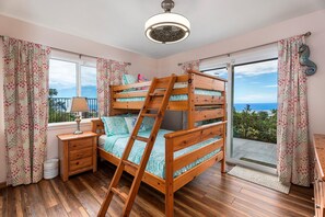 A bedroom with a bunk bed, wooden floor, a bedside table with a lamp, a window with curtains, and a view of the sea. - HALE LIO KAI: GORGEOUS HOME WITH POOL, HOT TUB & STUNNING OCEAN/SUNSET VIEWS (Kailua Kona)
