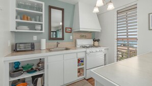 Une cuisine avec des meubles blancs, une cuisinière, un évier et une fenêtre avec des stores.
