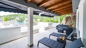 Ein überdachter Außenbereich mit modernen Möbeln, Blick auf einen Pool und Grünflächen sowie eine rustikale Steinmauer.