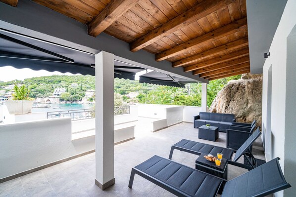 Un espace extérieur aménagé, abrité et doté de mobilier moderne, offrant une vue sur une piscine et sur la verdure, ainsi qu’un mur en pierre rustique.