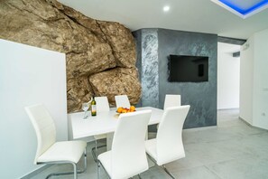 Un espace repas moderne avec une table blanche, des chaises blanches, un mur en pierres apparentes, un téléviseur à écran plat et un panier de fruits.