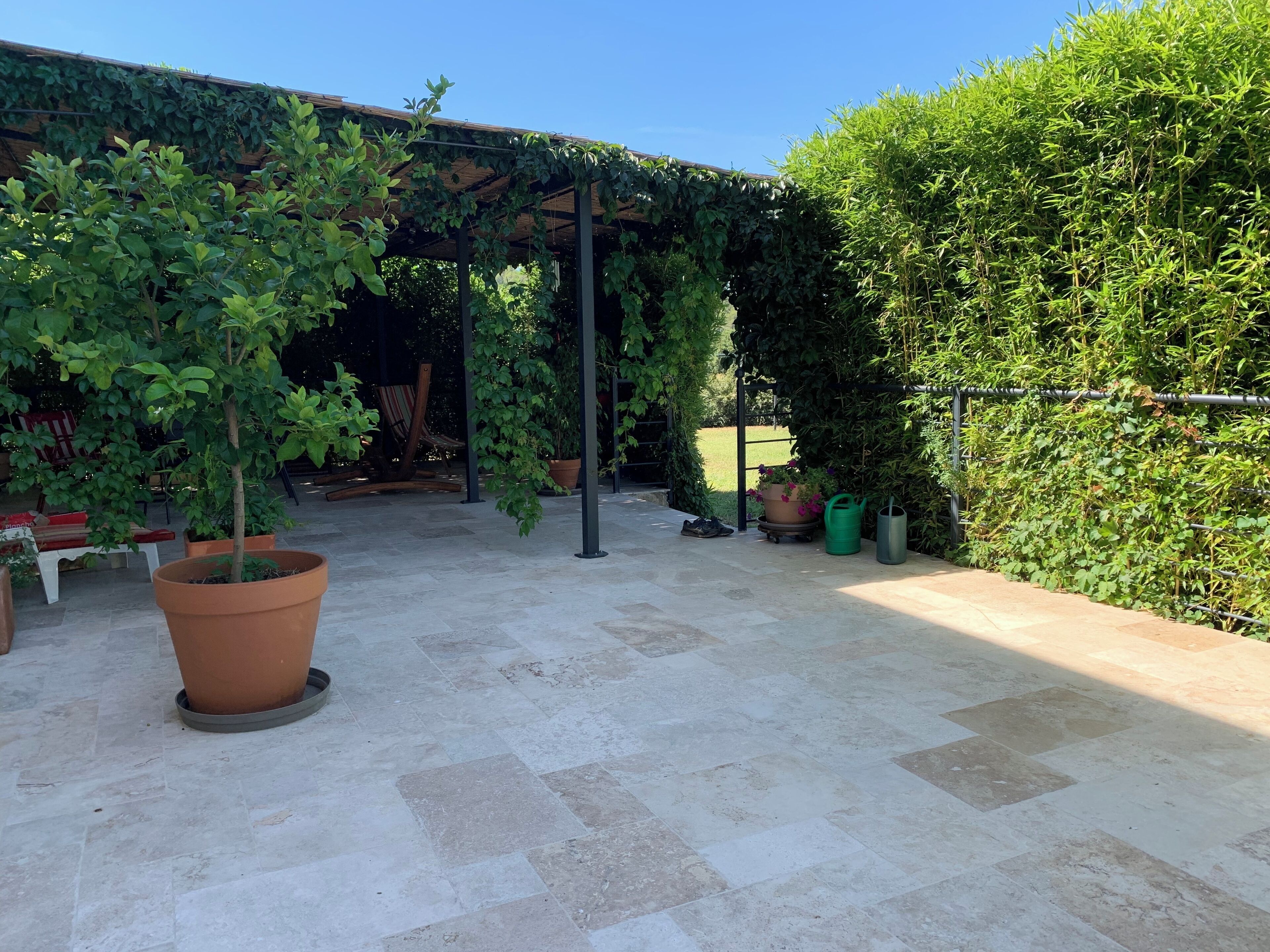 En täckt uteplats med en pergola, krukväxter och en sittgrupp med en bänk och stolar.