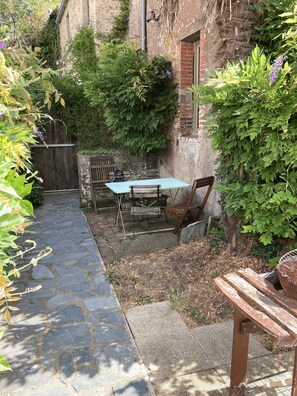 Eine terrasse mit Steinplatten, einem Tisch und Stühlen, umgeben von Grünpflanzen und einem rustikalen Gebäude.