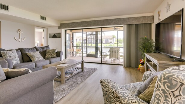A modern living room with a grey sofa, patterned armchair, and a flat-screen TV. - Covered Lanai & Waterfront View: Cape Coral Condo (Cape Coral)
