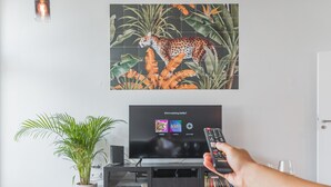 Ein Wohnzimmer mit Fernseher, der eine Netflix-Oberfläche zeigt, einer Zimmerpflanze und einem Glastisch.