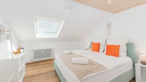Ein Schlafzimmer mit schrägem Dach, einem Dachfenster, einem Bett mit weißer und orangefarbener Bettwäsche, einem Traumfänger und einer weißen Heizung.
