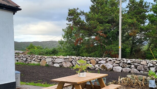 A wooden picnic table with benches, surrounded by gravel, with a stone wall and trees in the background.