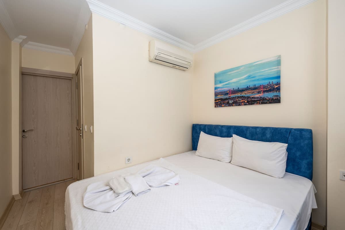 A hotel room with a bed, white linens, a blue headboard, a painting of a cityscape, and an air conditioning unit.
