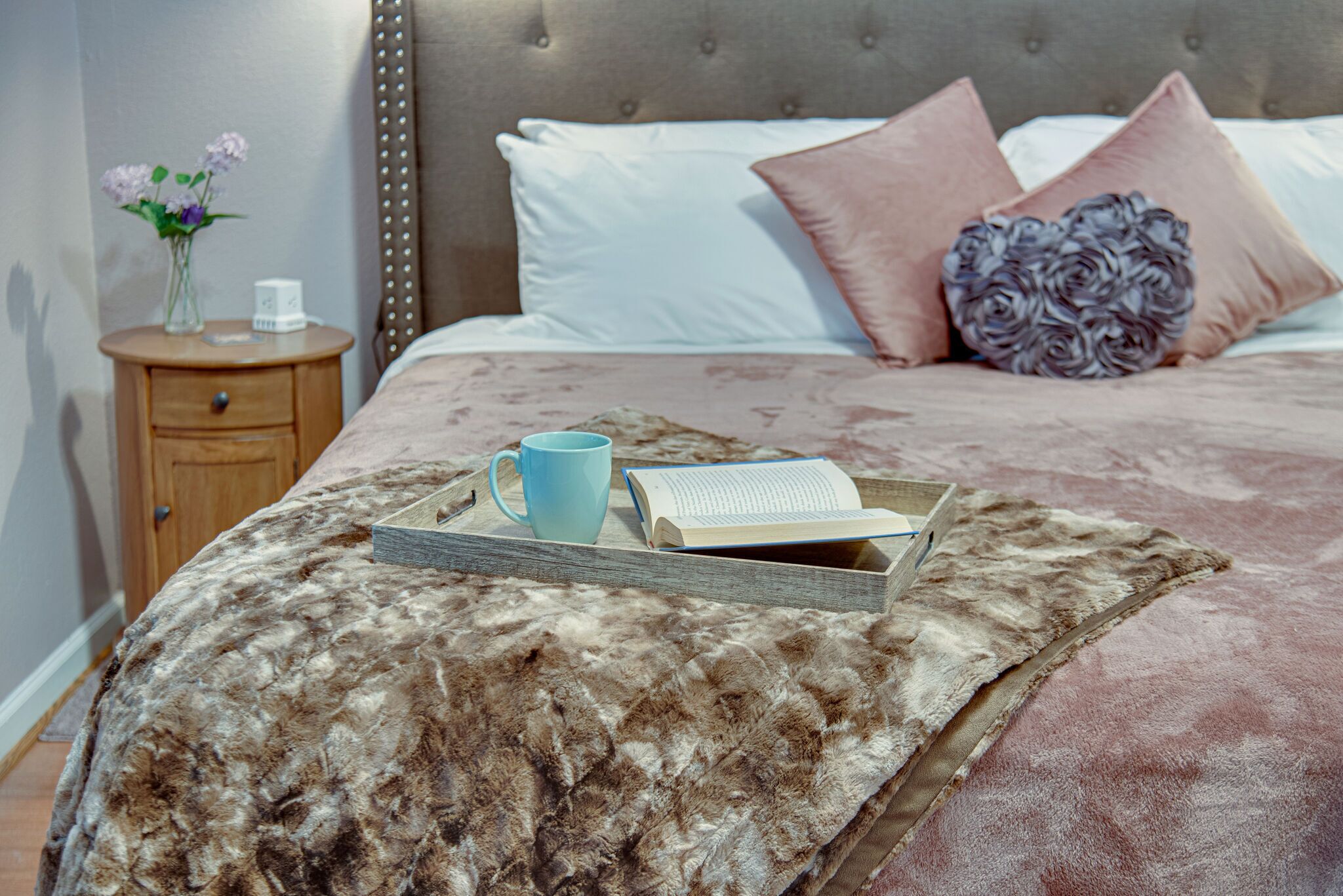 Una cama bien hecha con un edredón mullido, una mesita de noche con un florero y una bandeja con un libro y una taza.
