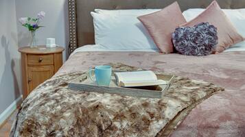 Una cama bien hecha con un edredón mullido, una mesita de noche con un florero y una bandeja con un libro y una taza.