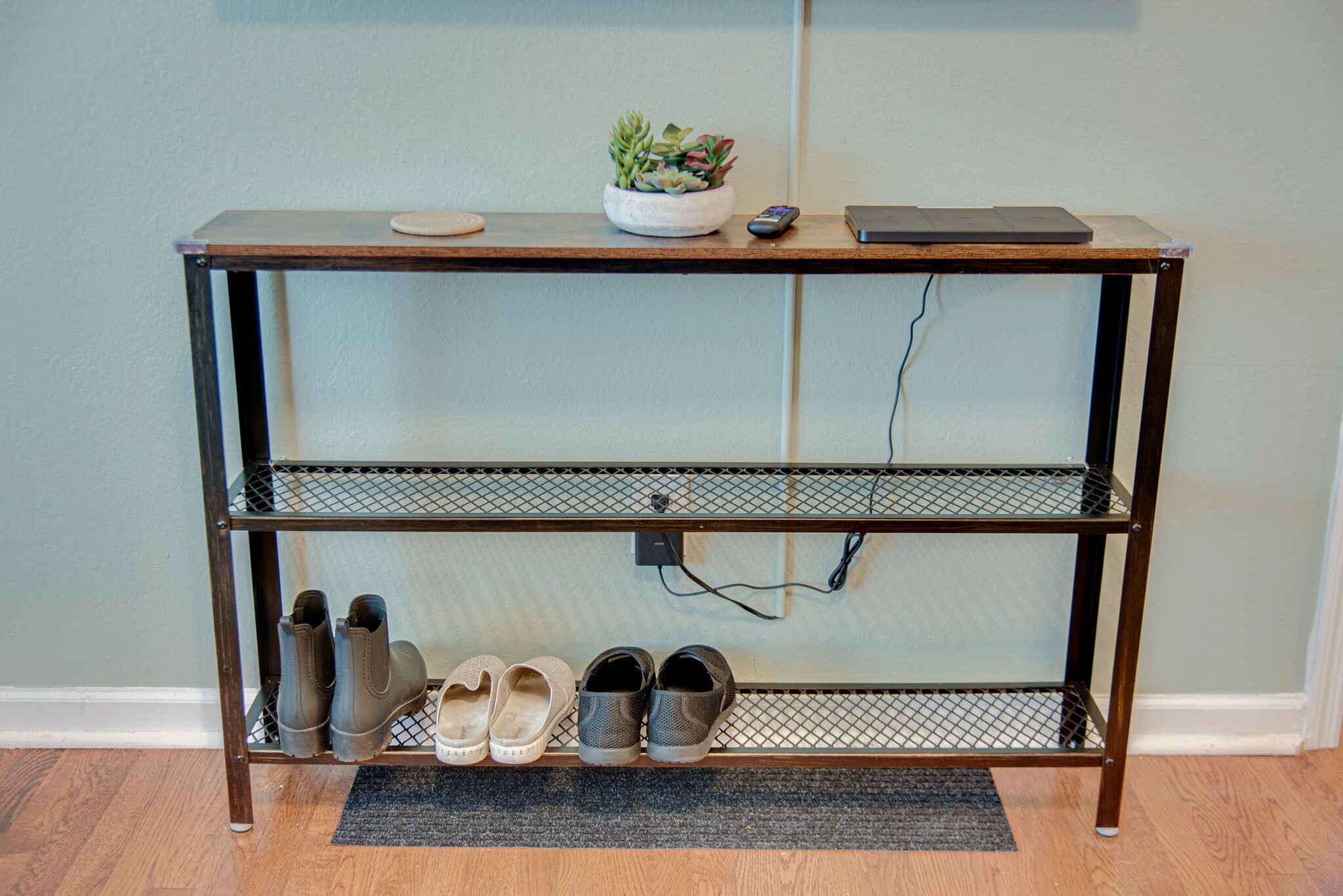 A metal wire shelf with three levels, holding shoes and a plant.