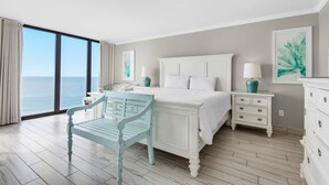 A bedroom with a large bed, a chair, a nightstand, and a dresser. There is a window with a view of the ocean.