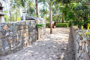 Un sentier pavé de pierres, bordé de murs en pierre, qui traverse une zone verdoyante et luxuriante, parsemée d’arbres et de plantes.