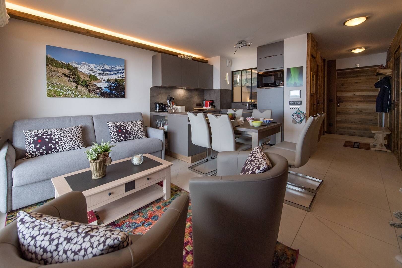 Un salon moderne avec un canapé gris, une table basse et un coin repas comprenant des chaises, et une cuisine en arrière-plan.
