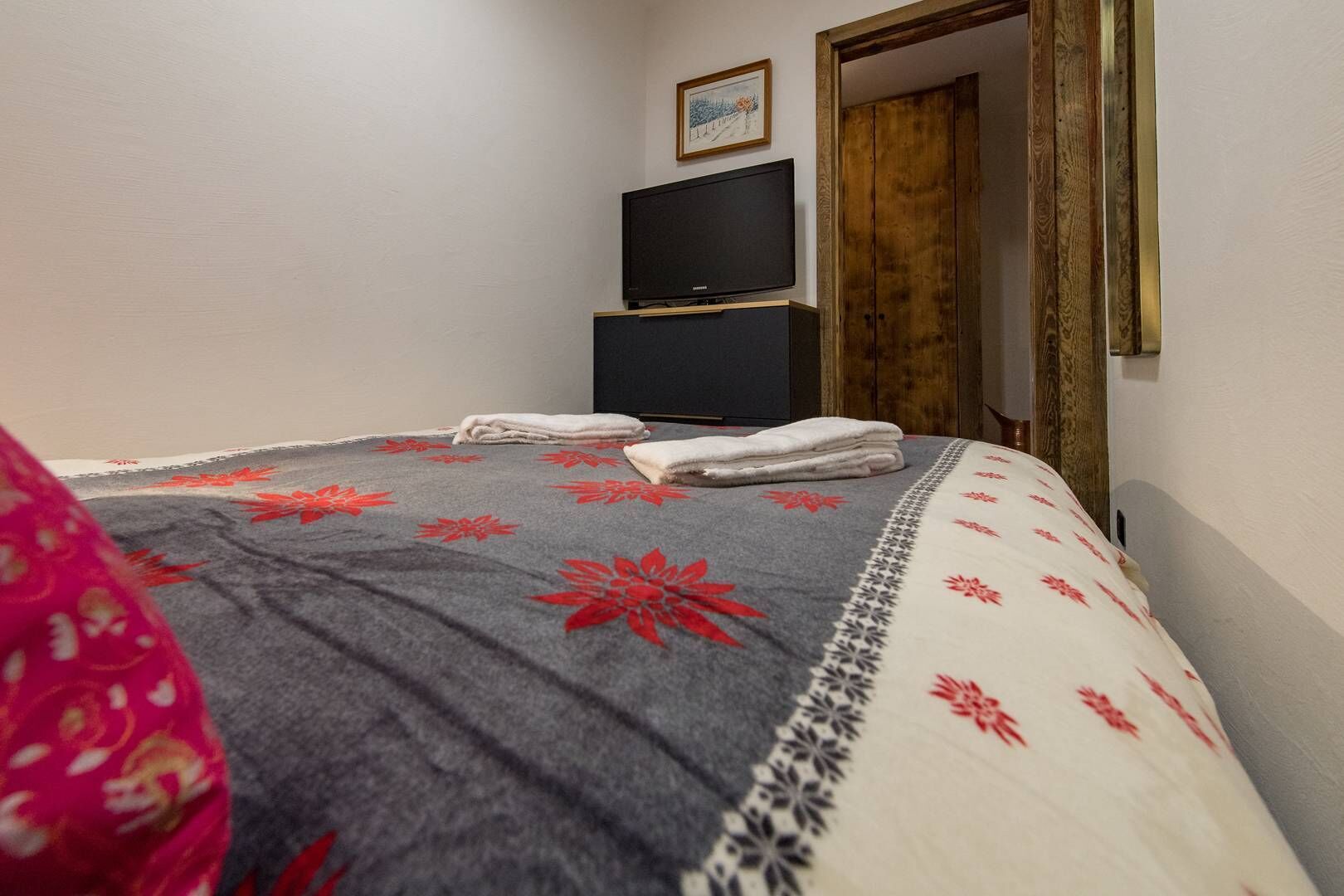 A neatly made bed with a patterned bedspread, a folded towel on the bed, and a television on a stand against the wall.