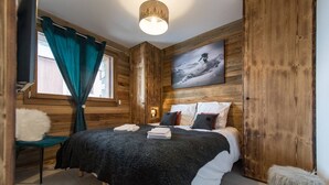 A wooden room with a bed, a window with curtains, a painting on the wall, and a small stool. - Olvera 203 - New apartment at the foot of the slopes (VALMEINIER)