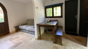 A room with a wooden table, a sofa, a window with a view of greenery, and a dark door. - The little House in Gigaro (La Croix-Valmer)