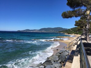 A coastal walkway with a rocky barrier, a clear blue sea, and distant hills.