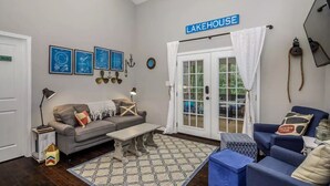 Uma sala de estar com um sofá cinzento, uma poltrona azul, uma mesa de centro e um tapete estampado. Há uma porta com a inscrição "Lakehouse" e uma janela com cortinas.