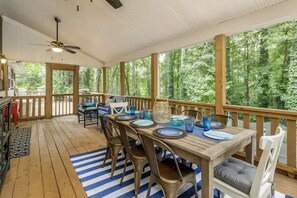 A covered outdoor dining area with a wooden table set for four, surrounded by chairs, and a view of a forested area through large windows.