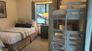 Een slaapkamer met een stapelbed, een eenpersoonsbed, een dressoir, een lamp en een raam met gordijnen.