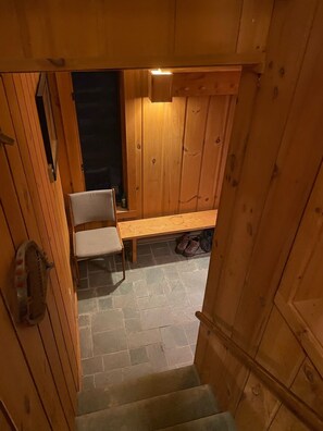 Interior de uma cabana de madeira com soalho em tijolos de pedra, um banco e uma cadeira.