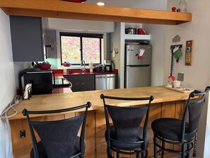 Une cuisine avec un îlot central en bois, deux chaises, un réfrigérateur et une fenêtre donnant sur un paysage aux feuillages colorés.