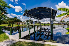 A covered outdoor area with a dock extending into a calm water body, surrounded by greenery and a clear sky.