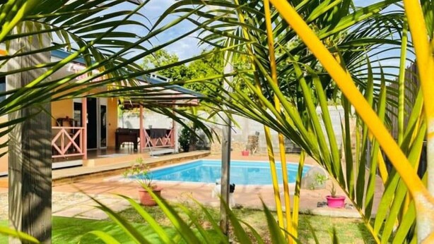 A swimming pool surrounded by lush greenery, a covered patio, and a building with a distinctive pink railing.