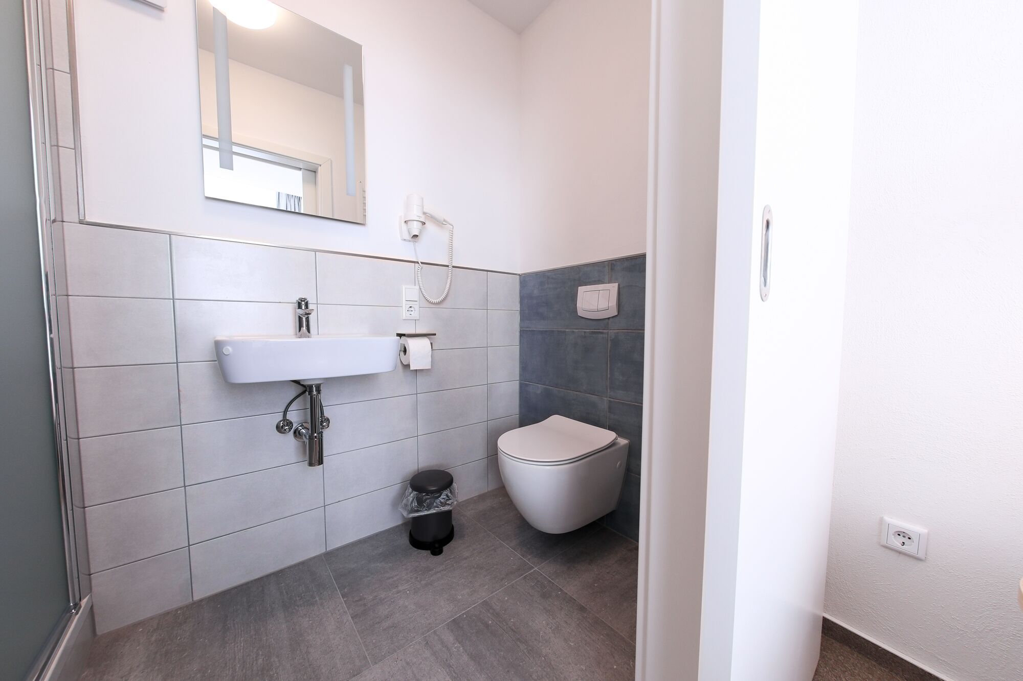 Un baño con lavamanos blanco, un espejo, un inodoro y un cubo de basura.
