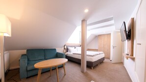 A modern, well-lit room with a sofa, a small round table, a bed, and a TV mounted on the wall. - 247 selfcheckin Schmiedestrasse (Leer)