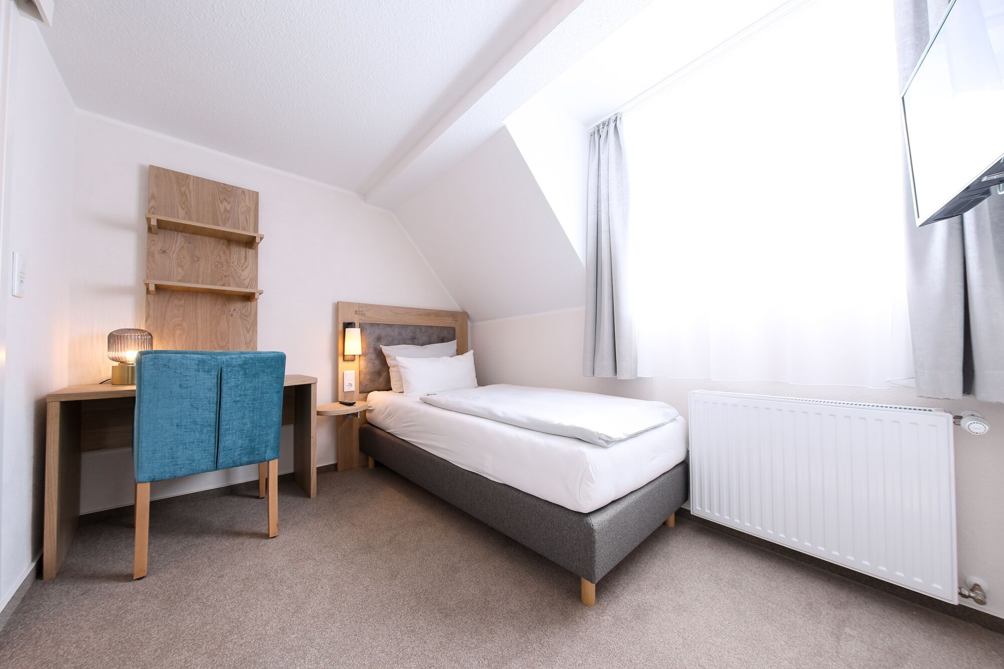 A bedroom with a bed, a desk, a chair, a nightstand, and a window with curtains.