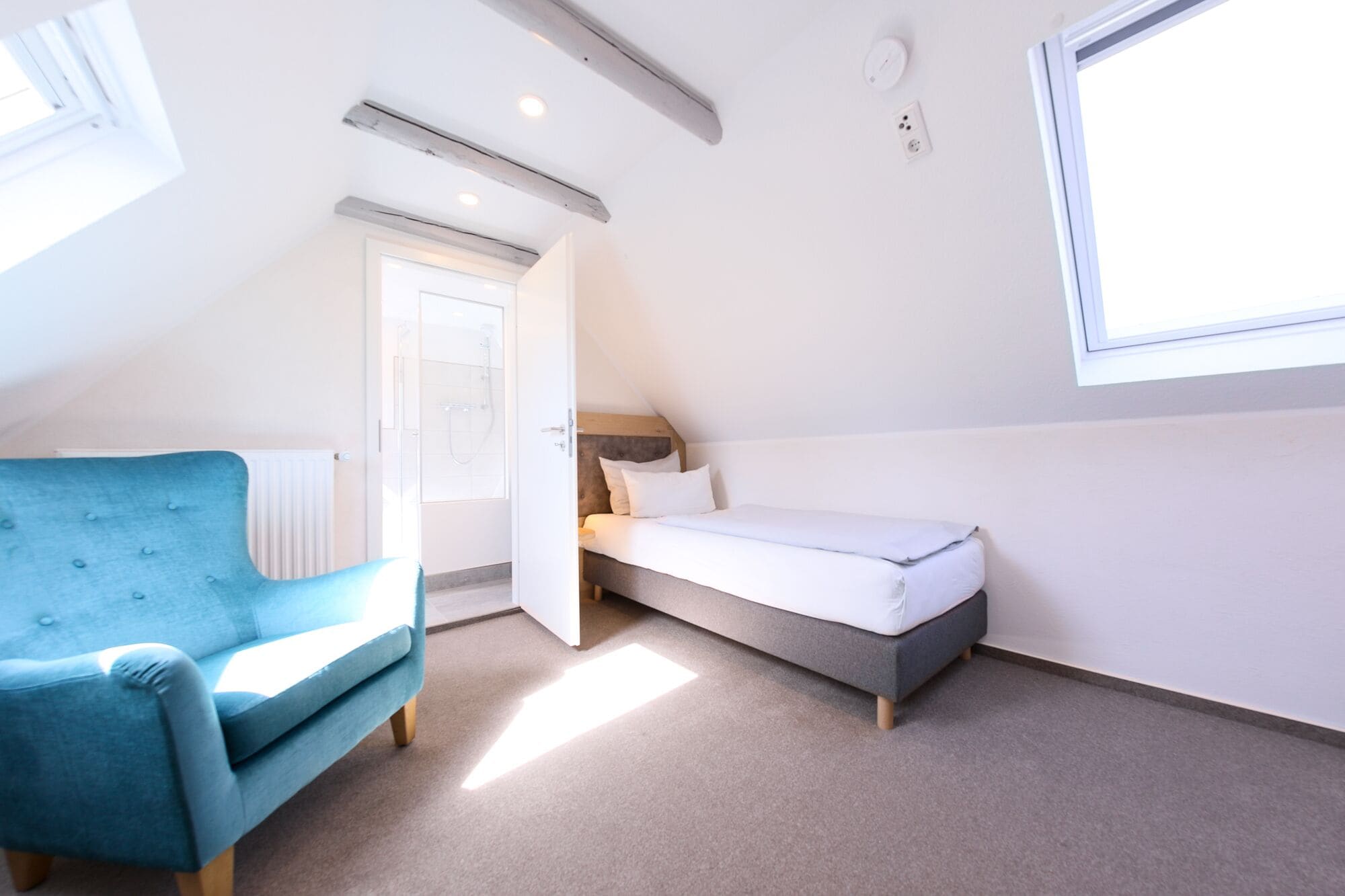 Una habitación pequeña y bien iluminada, con una cama individual, un sillón azul y una ventana que deja entrar luz natural.
