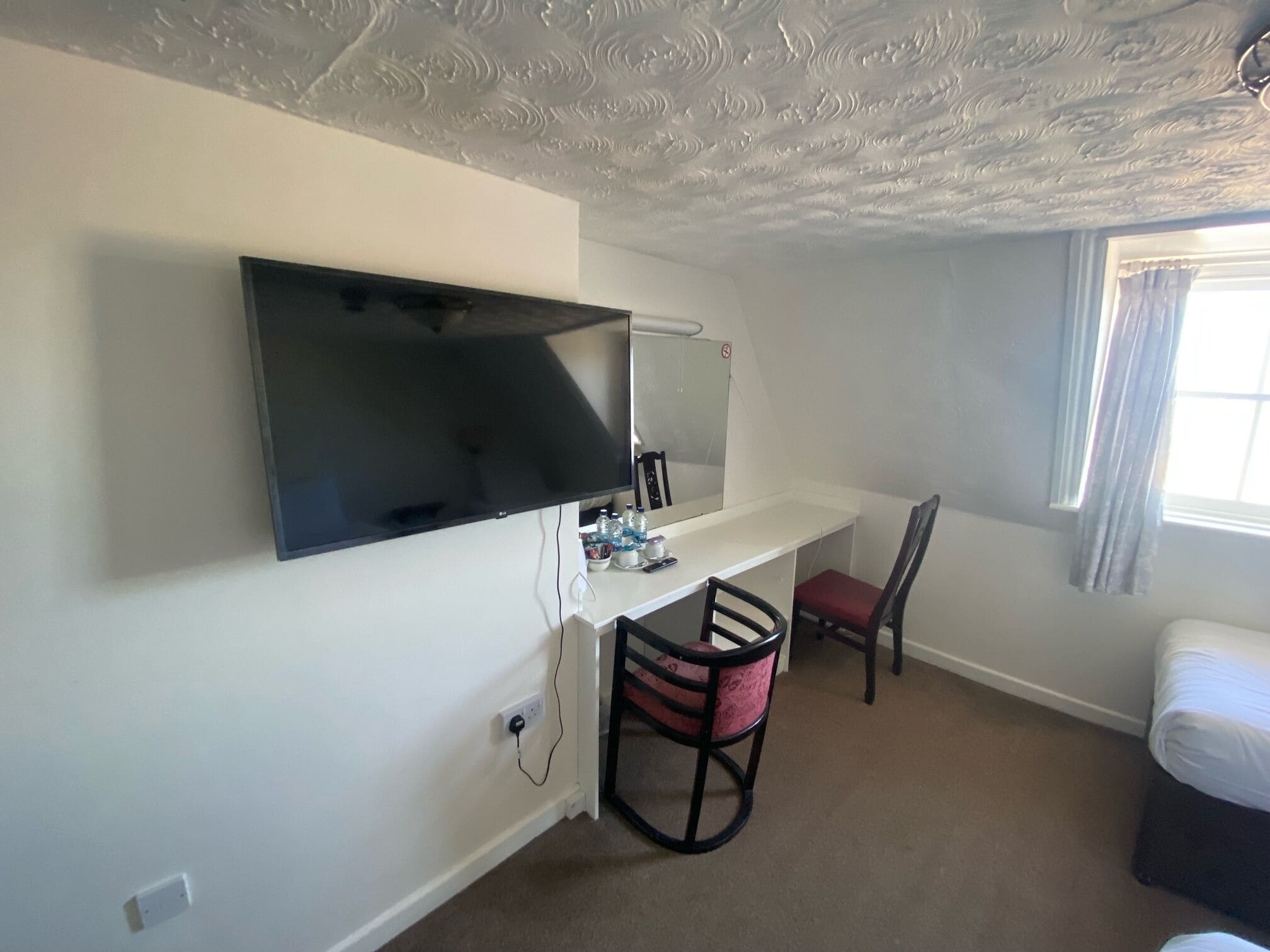 Habitación de hotel con un televisor de pantalla plana grande montado en la pared, un escritorio pequeño con una silla, una cama y una ventana con cortinas.