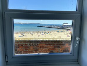 A view through a window of a beach with a brick wall, a pier, and a building in the background. - Fairhaven Hotel (Weymouth)