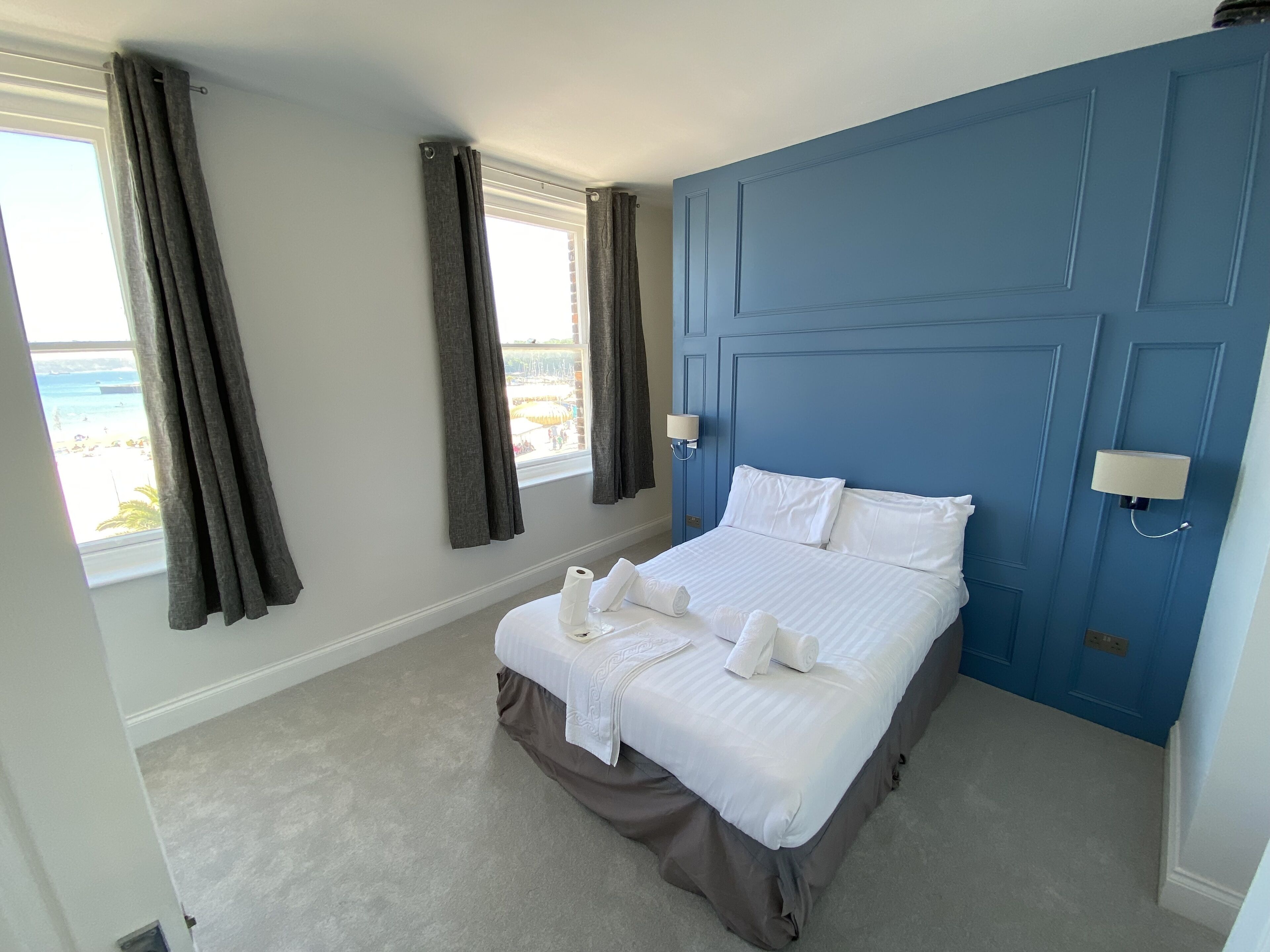 A hotel room with a bed, two windows with curtains, a blue accent wall, and a view of the beach.