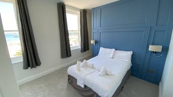 A hotel room with a bed, two windows with curtains, a blue accent wall, and a view of the beach.