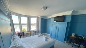 A hotel room with a large bed, a desk, a chair, and a TV. There is a window with a view of the beach. - Fairhaven Hotel (Weymouth)