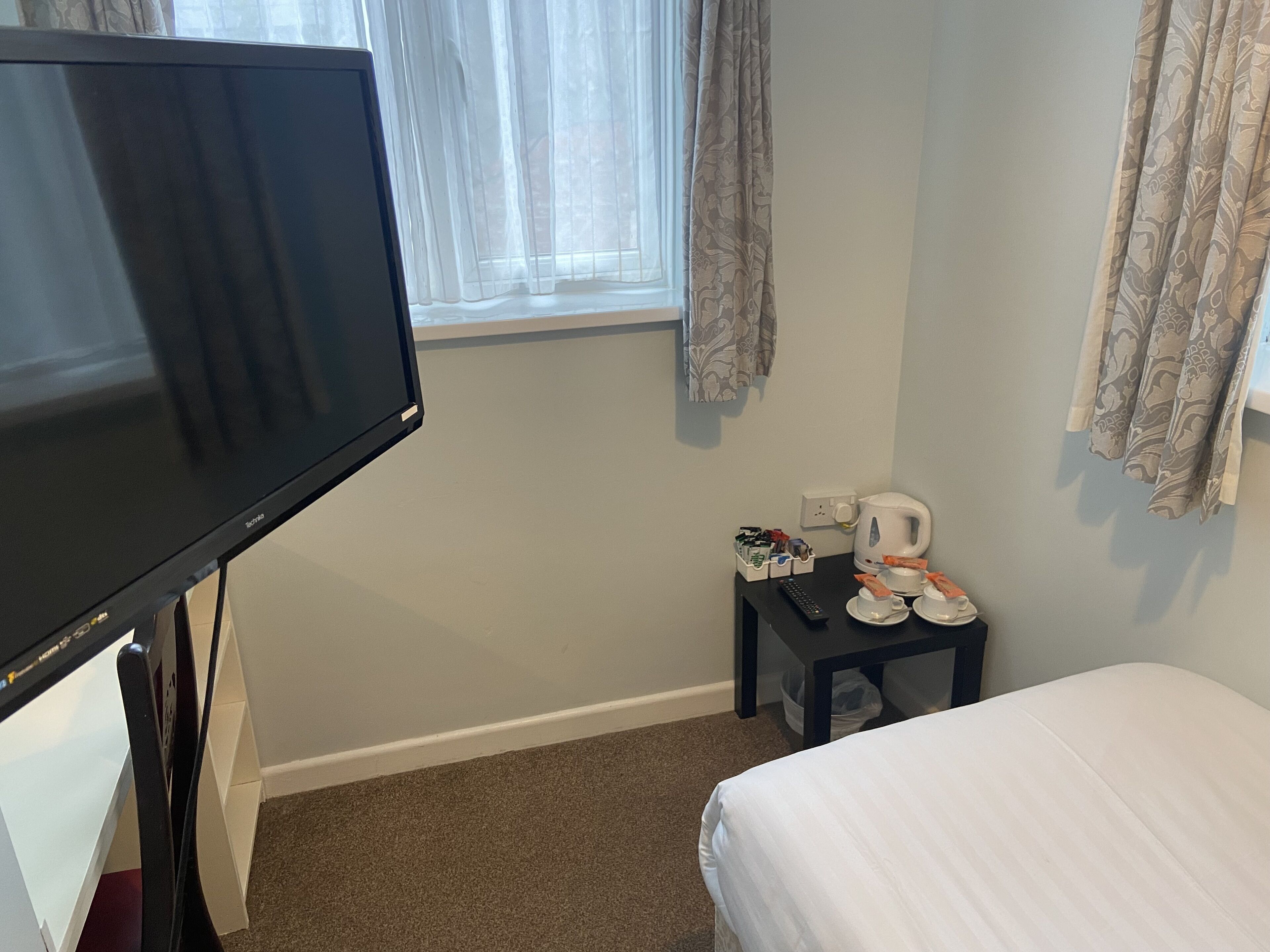 Habitación de hotel con cama, televisión, una mesita con hervidor, tazas y control remoto.
