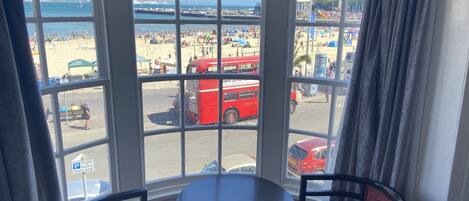A room with a bay window overlooking a beach. There is a round table and two chairs.