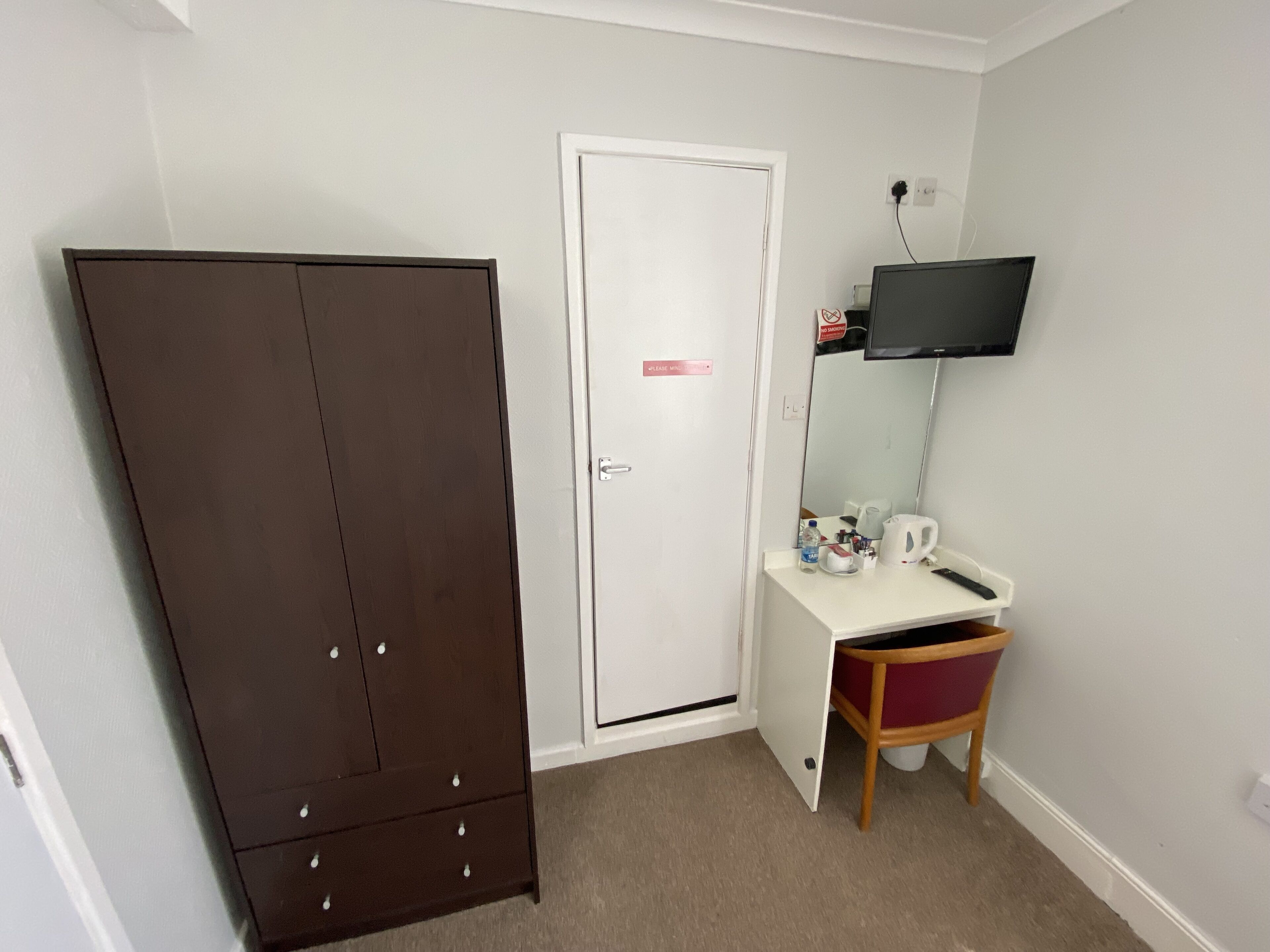 Una habitación con un armario marrón, un escritorio blanco con silla, un espejo y un televisor montado.