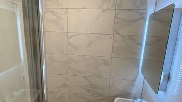 Een badkamer met een glazen douchecabine, een wit wastafelmeubel met een spiegel erboven en marmeren tegels op de muren.