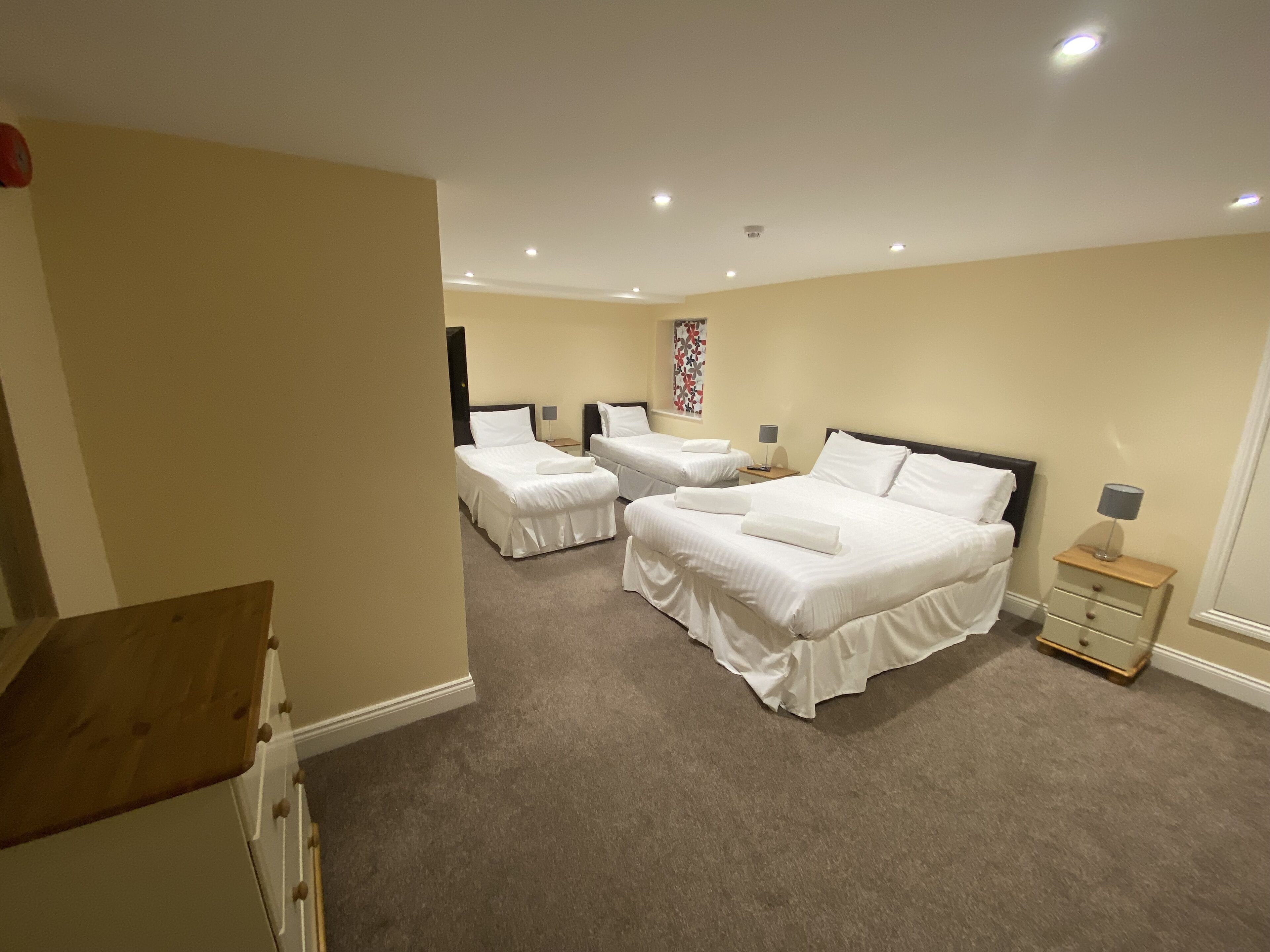 A hotel room with three beds, a wooden dresser, and a small bedside table with a lamp.