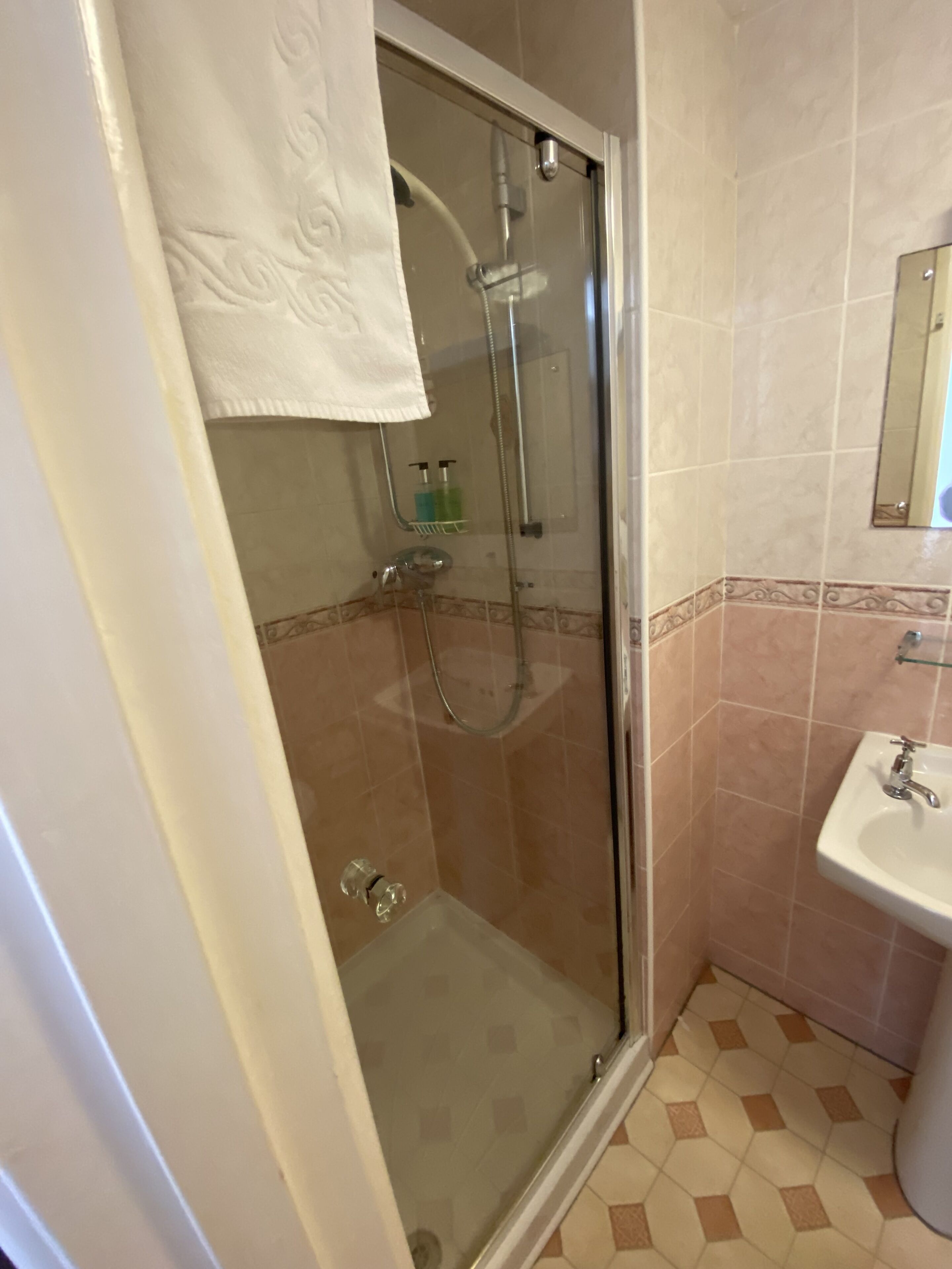 Cabina de ducha con paredes de vidrio, una toalla blanca colgada en la puerta, un cabezal de ducha de vidrio y un pequeño estante con artículos de tocador.