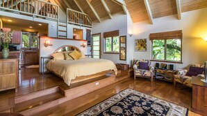 A spacious bedroom with a large bed, a wooden staircase, a sitting area with a chair and table, and a view of the outdoors through the windows. - Mahina Cottage, Mahina Cottage (Maui County)