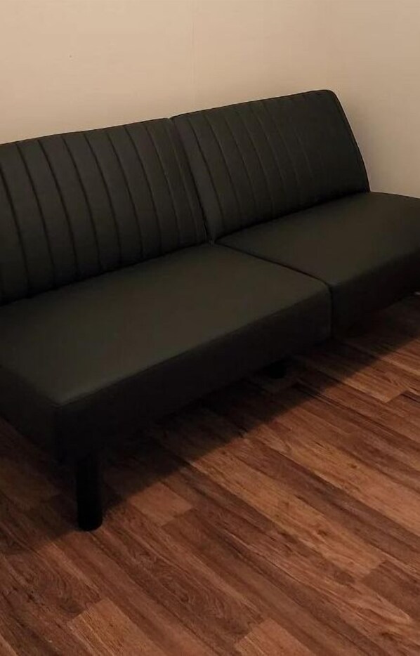 A dark green sofa with a ribbed backrest and armrests, positioned on a wooden floor against a plain wall.