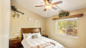 A bedroom with a bed, a ceiling fan, a window with a view of trees, and a nightstand with a lamp. - Mountain Retreat w/Foosball/Game Table! Fireplace~Large Deck~Near Park~Dogs OK! (Flagstaff)