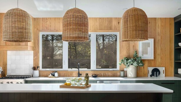 Une cuisine moderne dotée d’un grand îlot central, de murs recouverts de panneaux en bois et de trois suspensions lumineuses en fibres tressées.